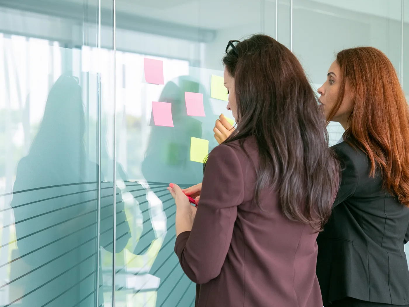 Two businesswomen arranging sticky notes on glass wall